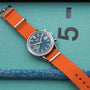 The TAG Heuer Limited Edition Carrera Skipper For HODINKEE, featuring an orange fabric strap and black dial, sits on a teal surface near a large 5 and blue-and-white braided rope—a bold pick for vintage chronograph enthusiasts.