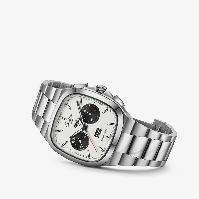 The Glashütte Original Seventies Chronograph Panorama Date is a silver wristwatch with a white face, black subdials, and a metal link band, displayed on a white background.