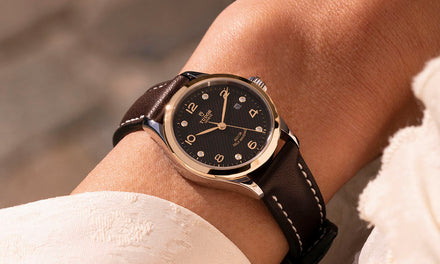 A close up shot of a person's wrist with the edge of a white shirt and a Tudor watch with a black textured dial and a brown leather strap