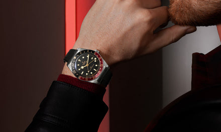 A man's chin rests on his closed fist, on his wrist is a Tudor watch with a black dial and a black and red two-tone bezel
