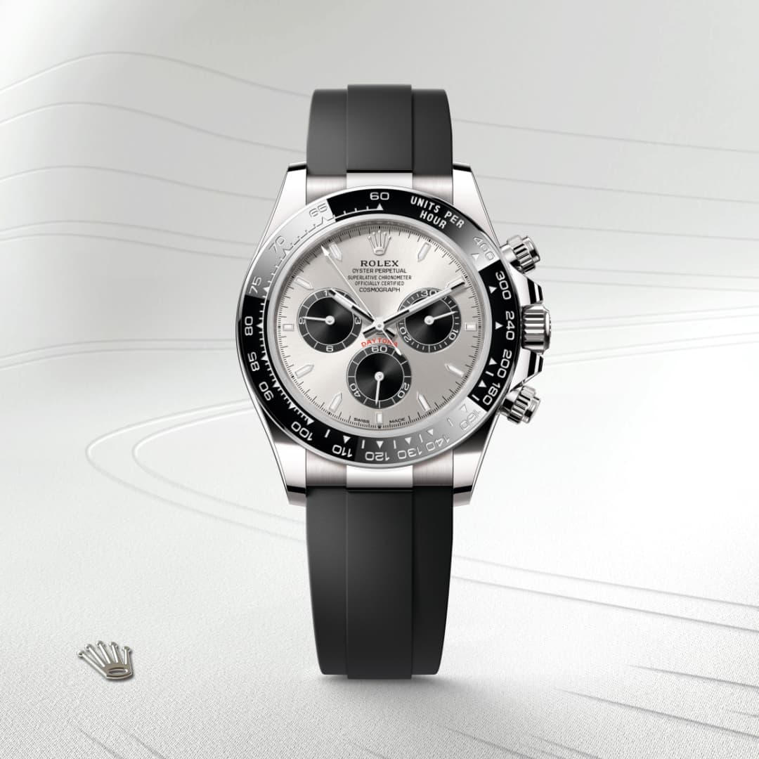 A silver Rolex chronograph watch with a black bezel and black rubber strap on a light textured background.