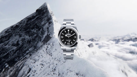 A silver wristwatch with a black face floats in front of a snow-covered mountain peak.
