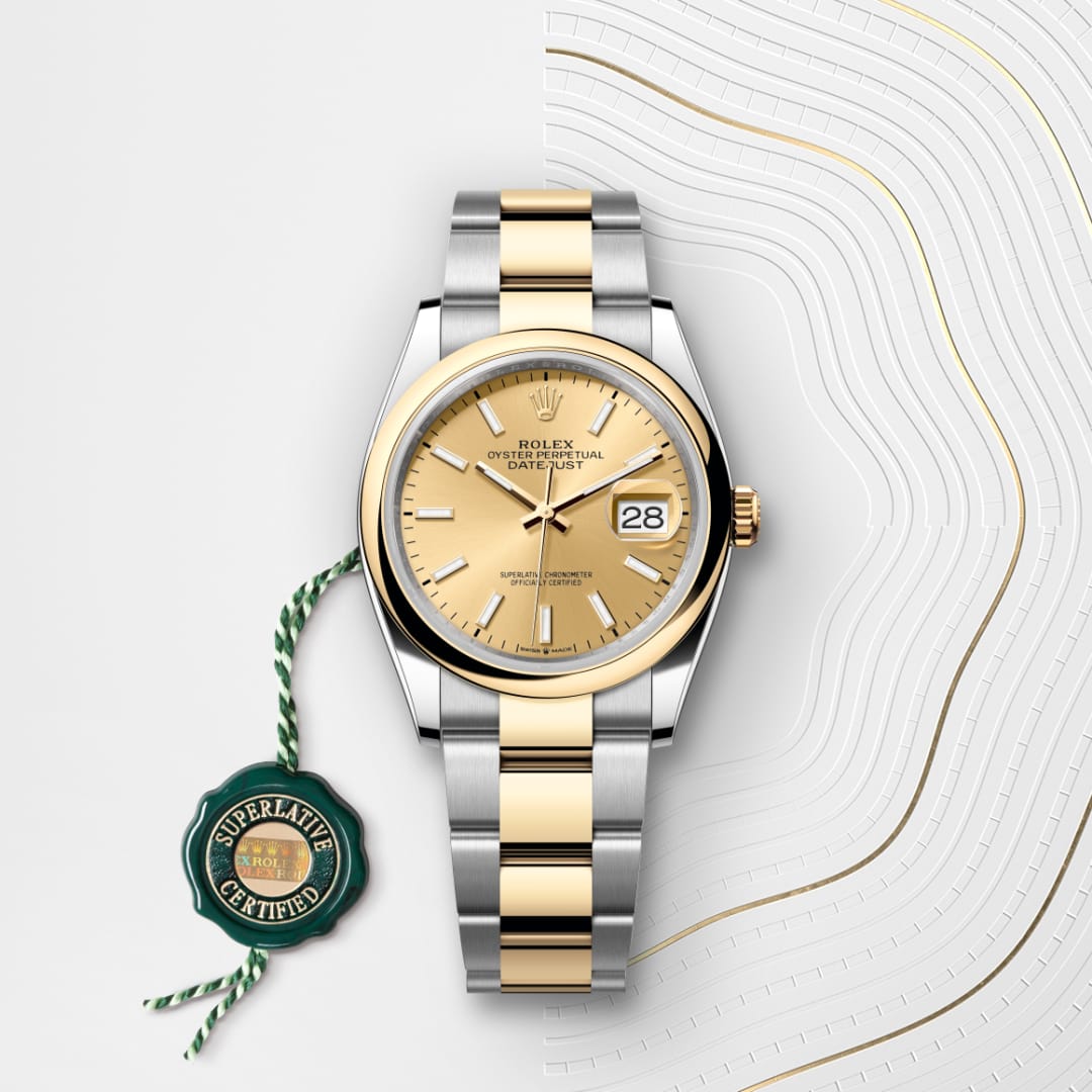A two-tone Rolex Datejust watch with a gold dial and date display, beside a “Superlative Certified” green tag.