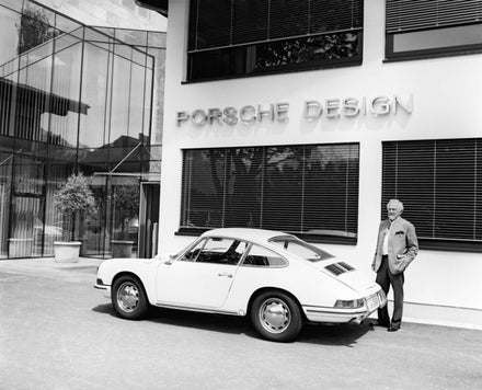 An older man stands by a classic white Porsche car in front of a building labeled Porsche Design.