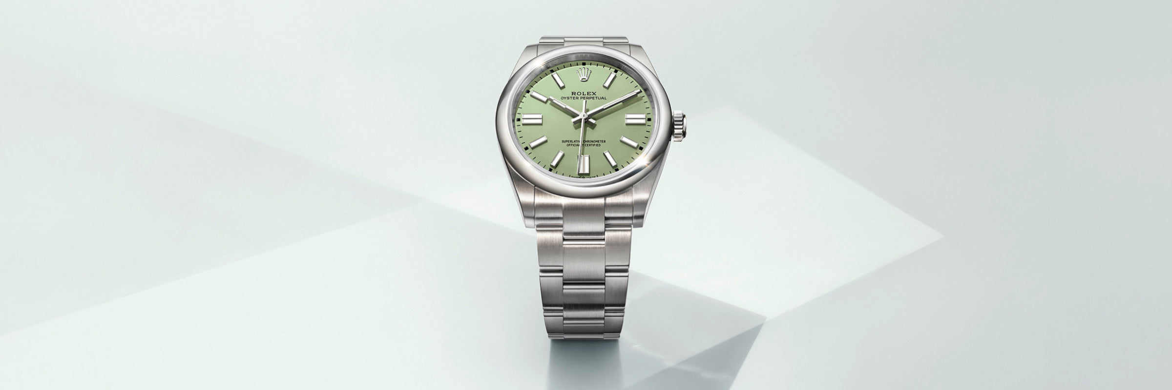 A silver Rolex wristwatch with a light green face displayed on a light, geometric background.