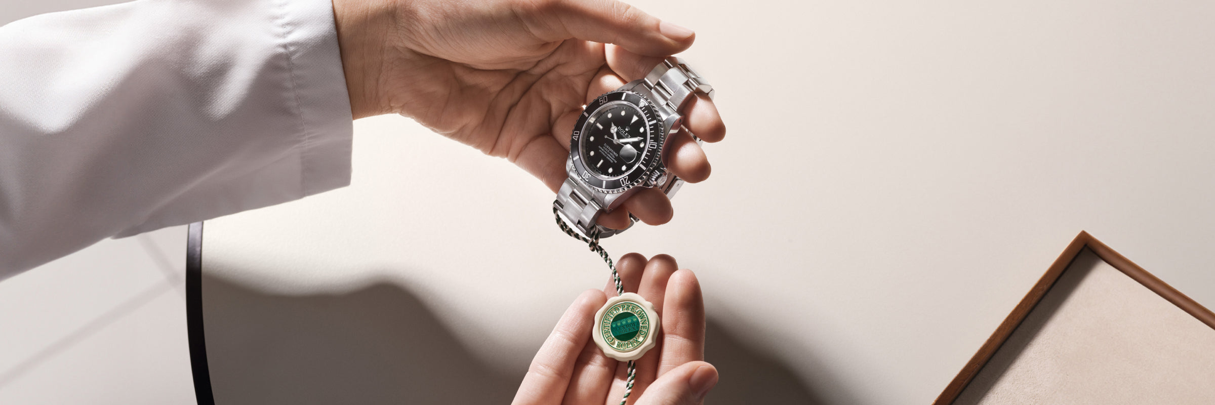 A person holds a luxury wristwatch and a green seal tag above a beige surface.