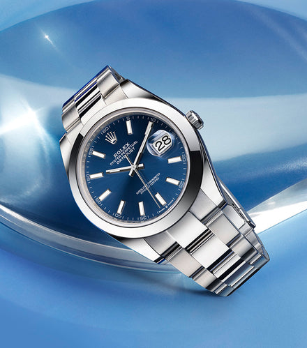 A silver Rolex watch with a blue dial and date display, resting on a glossy blue surface.