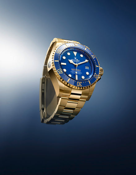 Gold Rolex wristwatch with a blue face and metal band, displayed on a gradient blue background.