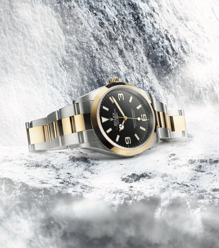 A two-tone Rolex watch with a black dial is displayed against an icy, waterfall-like background.