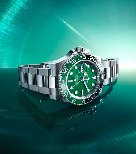A stainless steel luxury watch with a green dial and bezel displayed on a reflective teal surface.