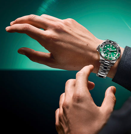 A person wearing a silver watch with a green face, adjusting it on their wrist against a green background.