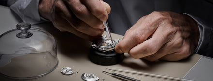 A watchmaker uses tools to carefully assemble the intricate parts of a mechanical watch on a workbench.