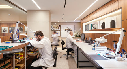 Watchmakers in white coats work at benches and desks in a bright, modern watch repair workshop.