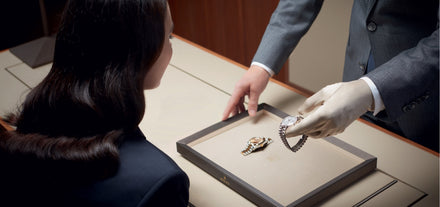 A salesperson in gloves shows a luxury watch to a woman seated at a display counter.