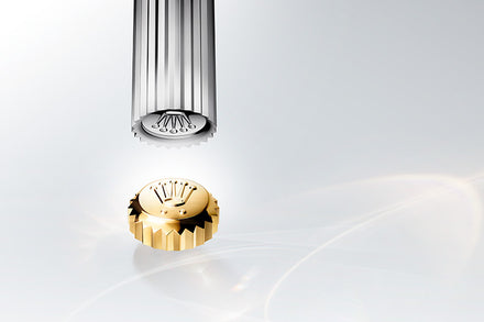 A gold Rolex crown with logo below a silver tool on a light, reflective background.
