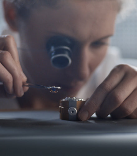 A person uses tweezers and a magnifying lens to assemble small parts, possibly working on a watch.