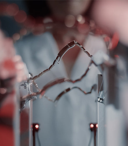 A person in a lab coat stands behind intricate metal wires and red lights in a scientific setting.
