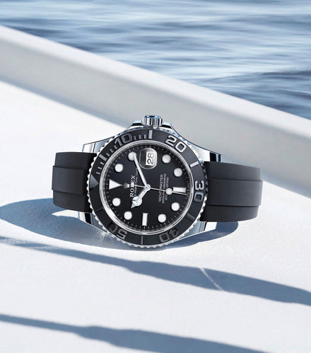 A black Rolex watch with a rubber strap rests on a white surface near water, casting a shadow.