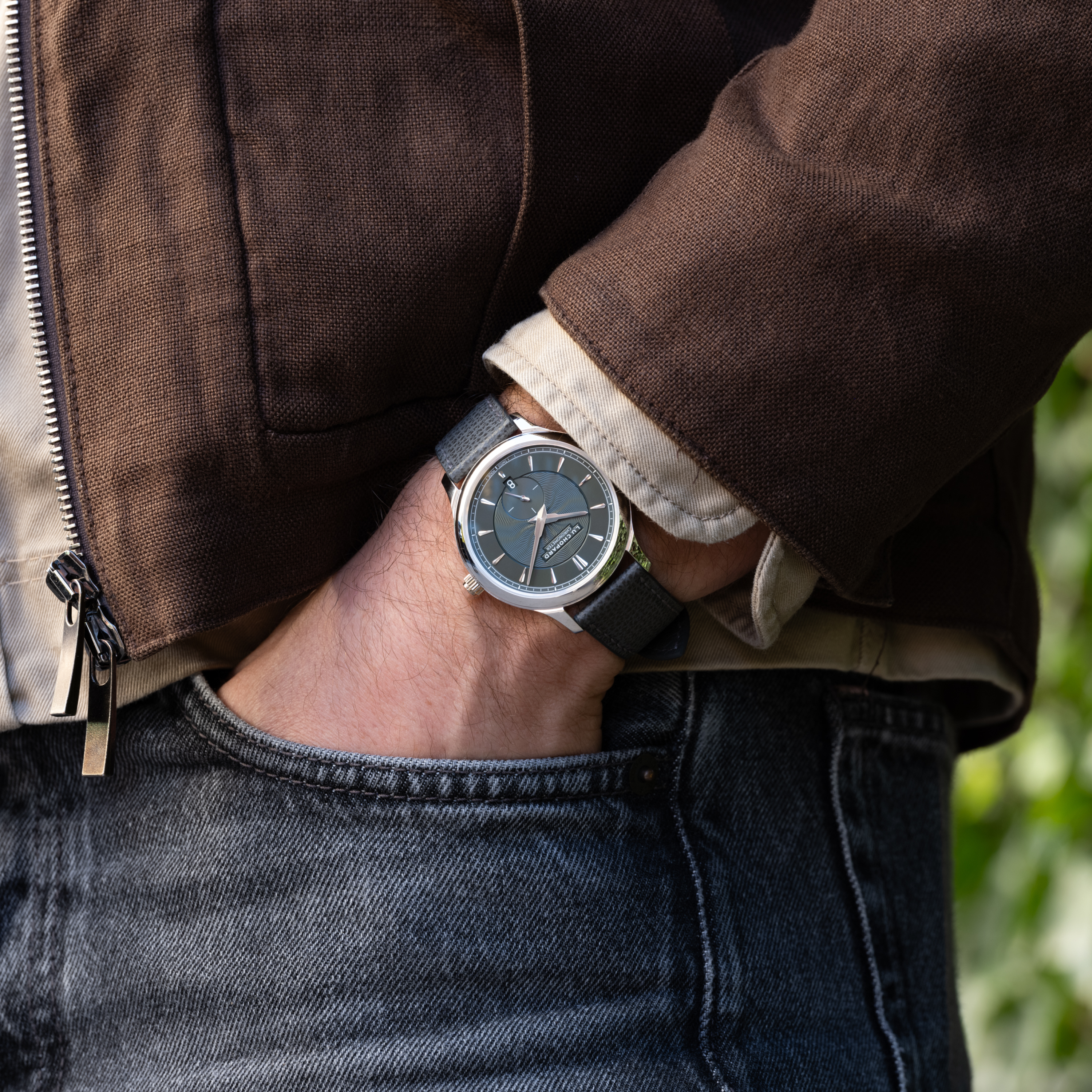 A person in a brown jacket and gray jeans, hand in pocket, wears a Chopard L.U.C XPS 1860 Officer Limited Edition For Hodinkee on a black strap.