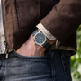 A person in a brown jacket and gray jeans, hand in pocket, wears a Chopard L.U.C XPS 1860 Officer Limited Edition For Hodinkee on a black strap.