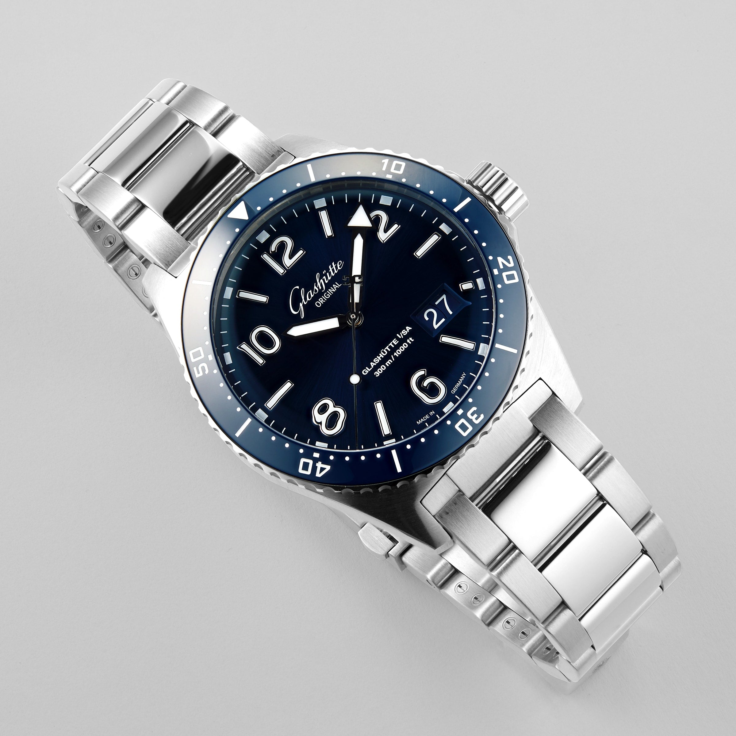 The Glashütte Original SeaQ Panorama Date is a stainless steel wristwatch with a dark blue dial, white hour markers, a date window at 3 o’clock, and a rotating bezel, shown against a light background.