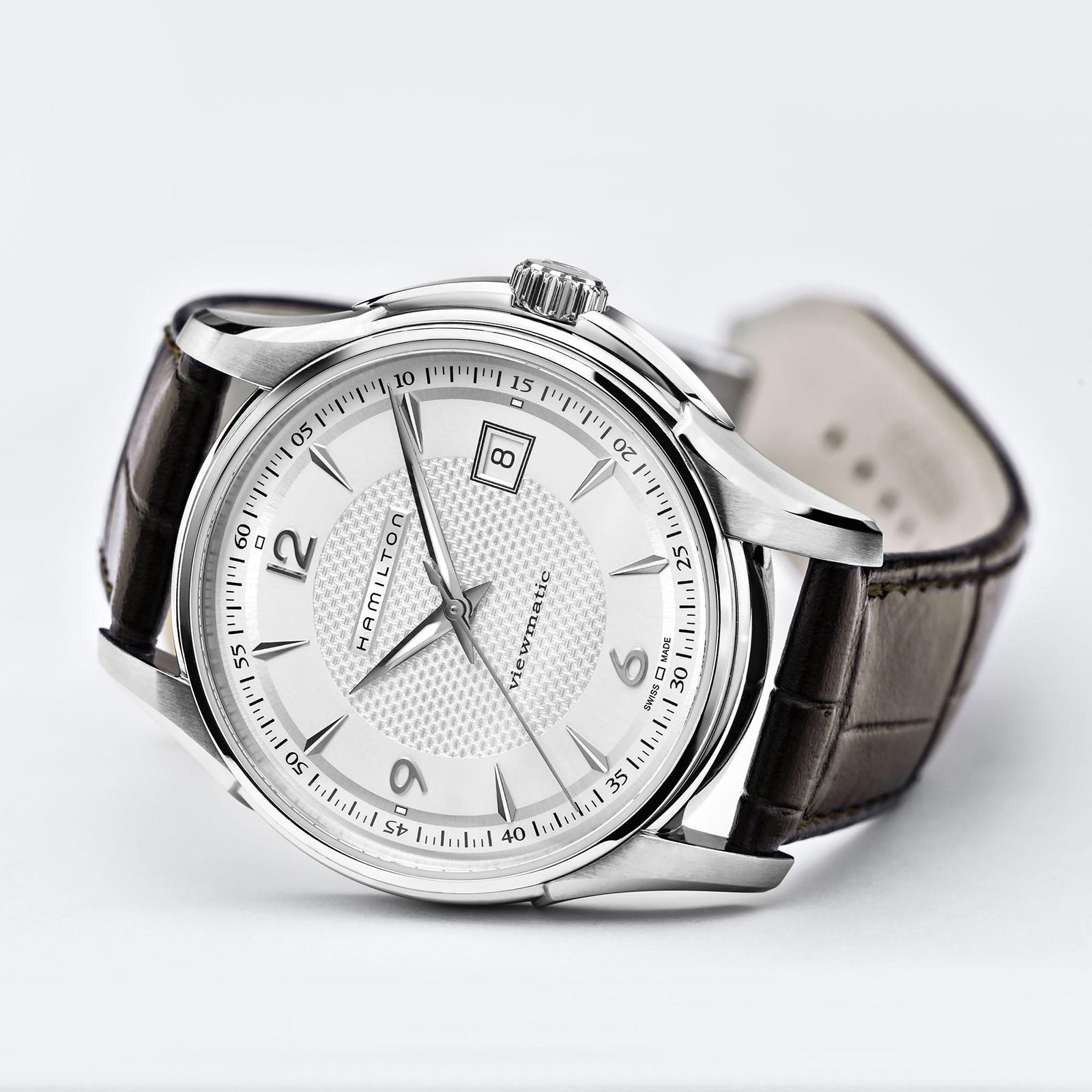 The Hamilton Jazzmaster Viewmatic Auto features a silver case, white dial with a date display at 3 o’clock, leather strap, and automatic movement—an elegant example of contemporary design—shown on a light background.