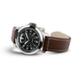 A Hamilton Khaki Field King Auto watch with a black dial, white numerals, and a brown leather strap with white stitching—featuring crown protectors—is displayed at an angle.