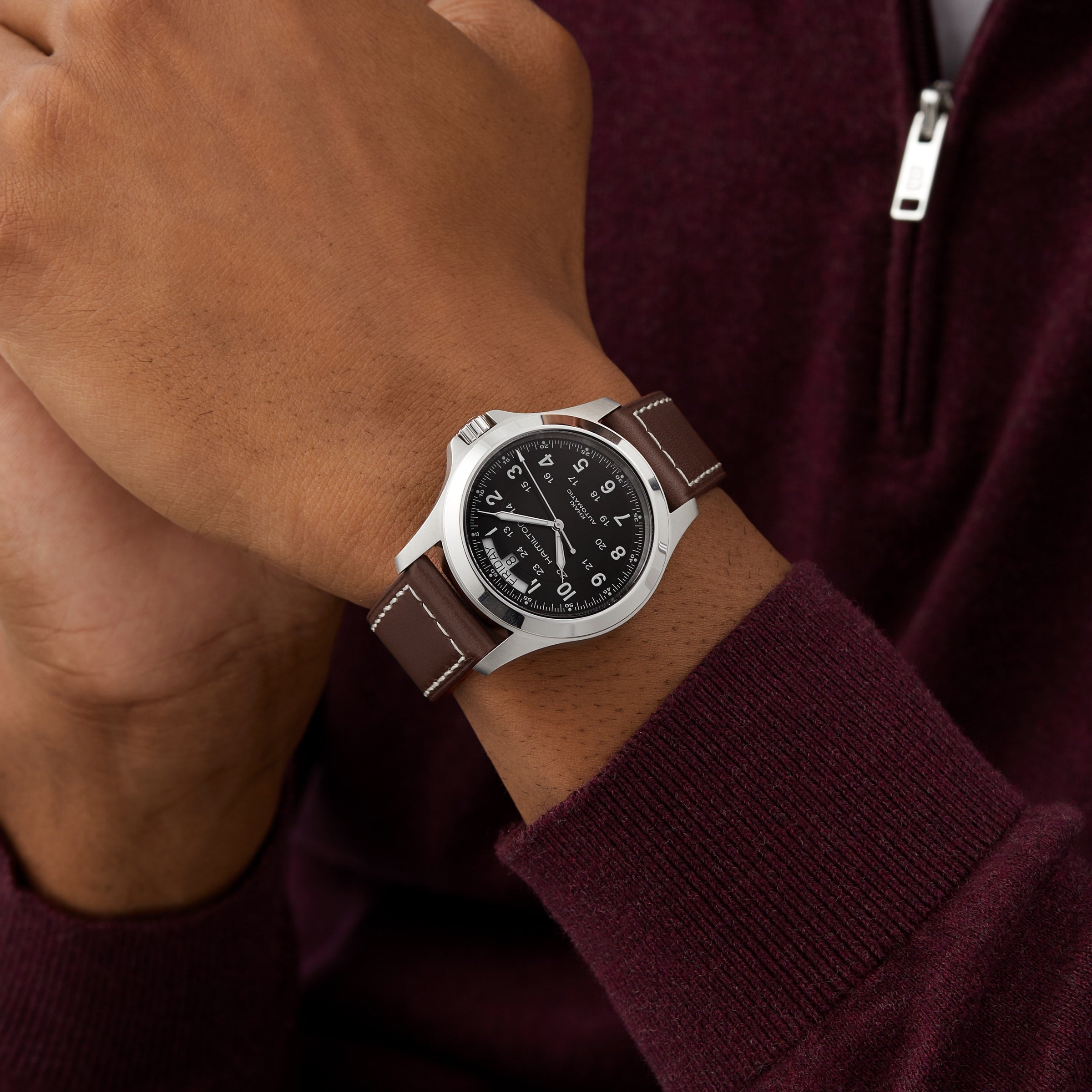 A person in a burgundy zip-up sweater wears a Hamilton Khaki Field King Auto watch, featuring a black dial with military time and a brown leather strap.
