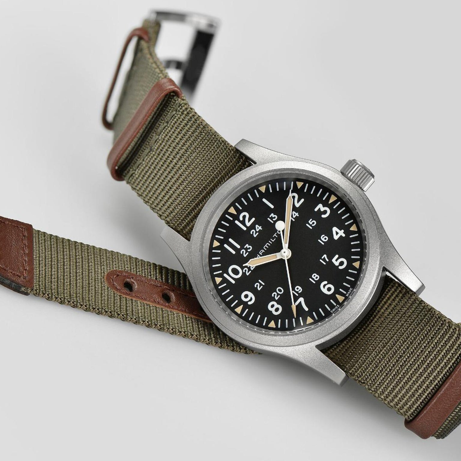The Hamilton Khaki Field Mechanical 38mm features a black dial with 12- and 24-hour markers, a stainless steel case, and an olive green NATO strap enhanced with brown leather accents.