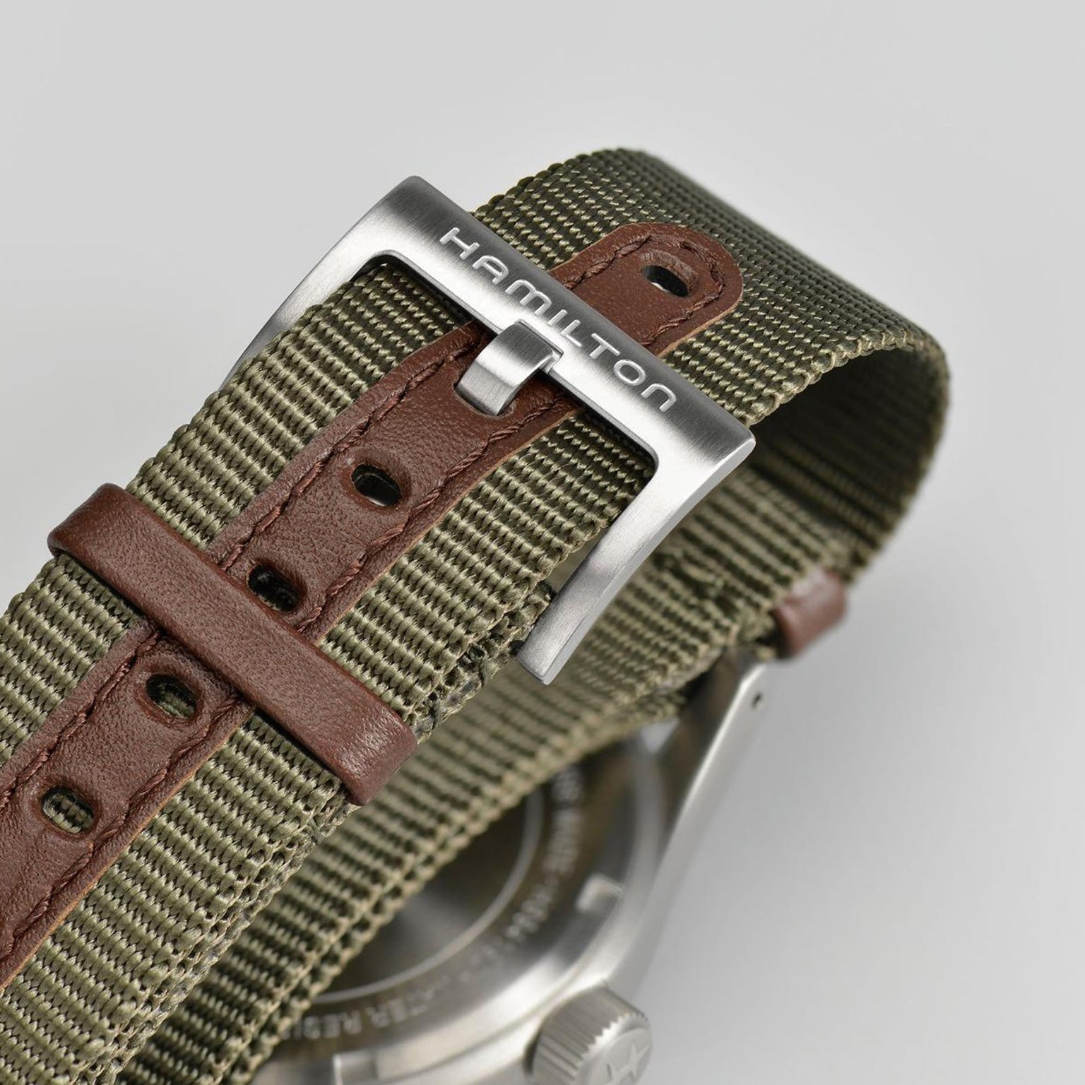 Close-up of a green fabric and brown leather NATO strap with a silver Hamilton-engraved buckle, attached to the stainless steel case of the Hamilton Khaki Field Mechanical 38mm watch.