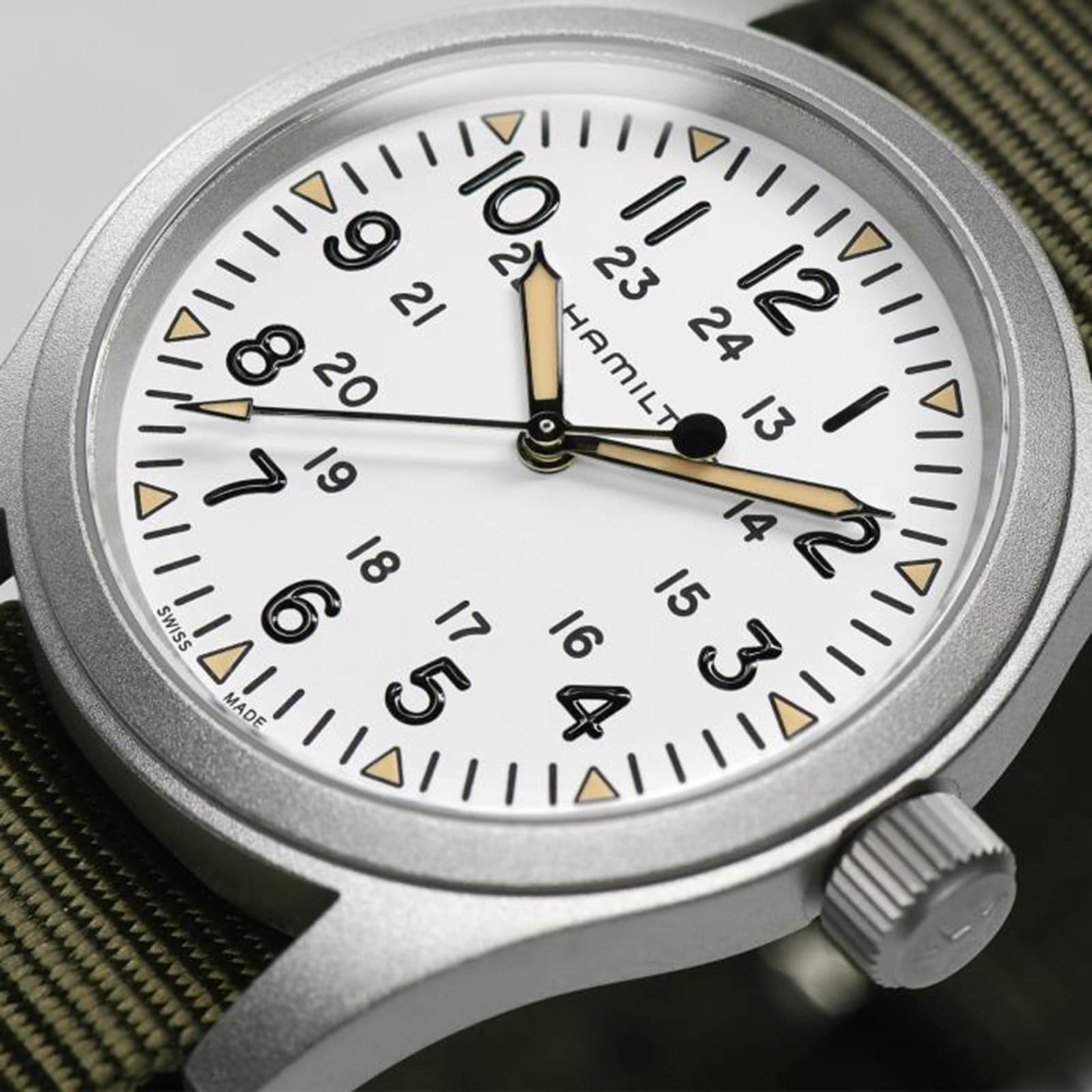 A close-up of the Hamilton Khaki Field Mechanical wristwatch, featuring a white dial, black numerals, and an olive green fabric strap.