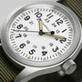 A close-up of the Hamilton Khaki Field Mechanical wristwatch, featuring a white dial, black numerals, and an olive green fabric strap.