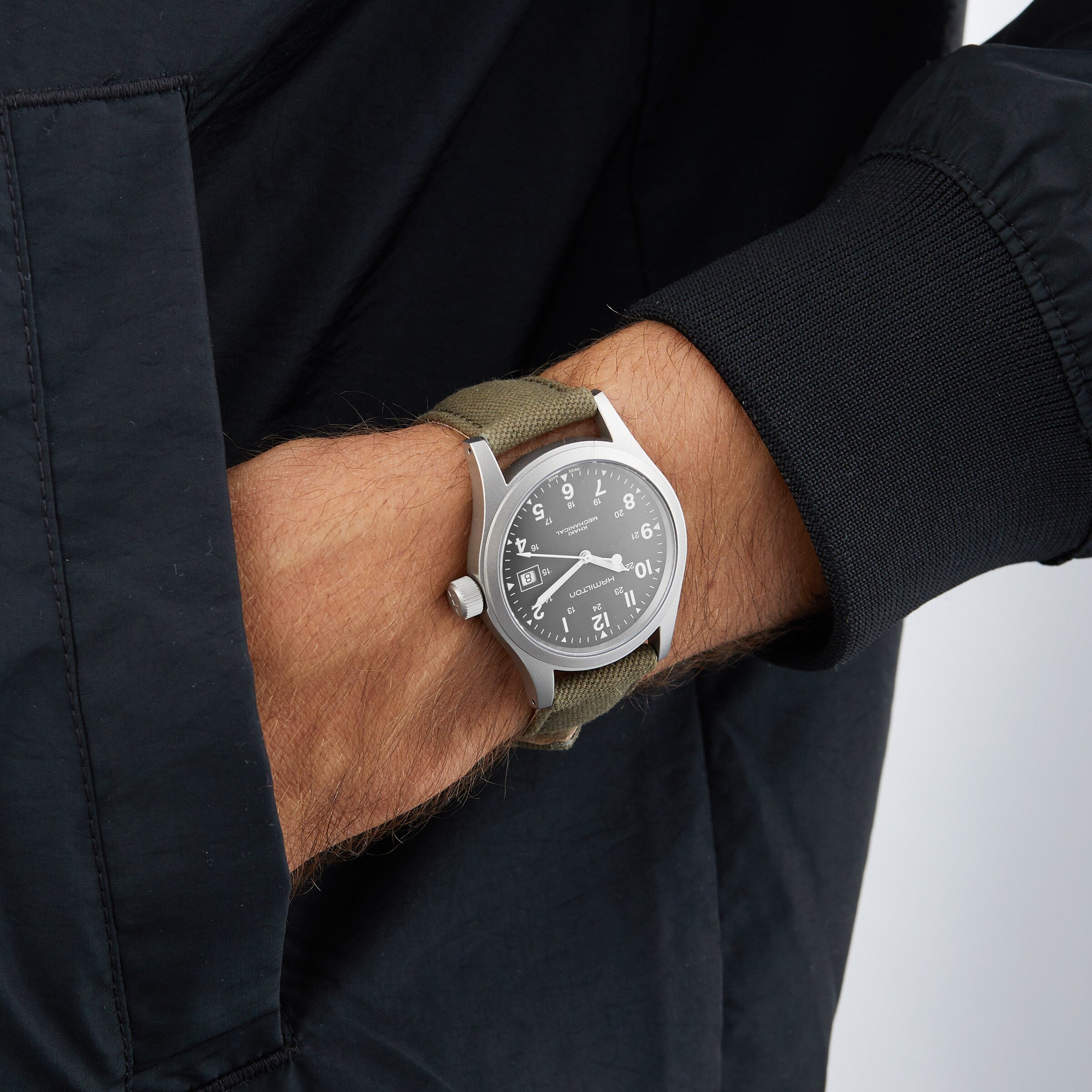 A person wearing a black jacket and a Hamilton Khaki Field Mechanical watch with a green strap on their left wrist.