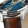 Close-up of the Hamilton Jazzmaster Open Heart Auto watch, featuring a smoked blue dial with visible movement, silver case, and brown leather strap in a modern style.