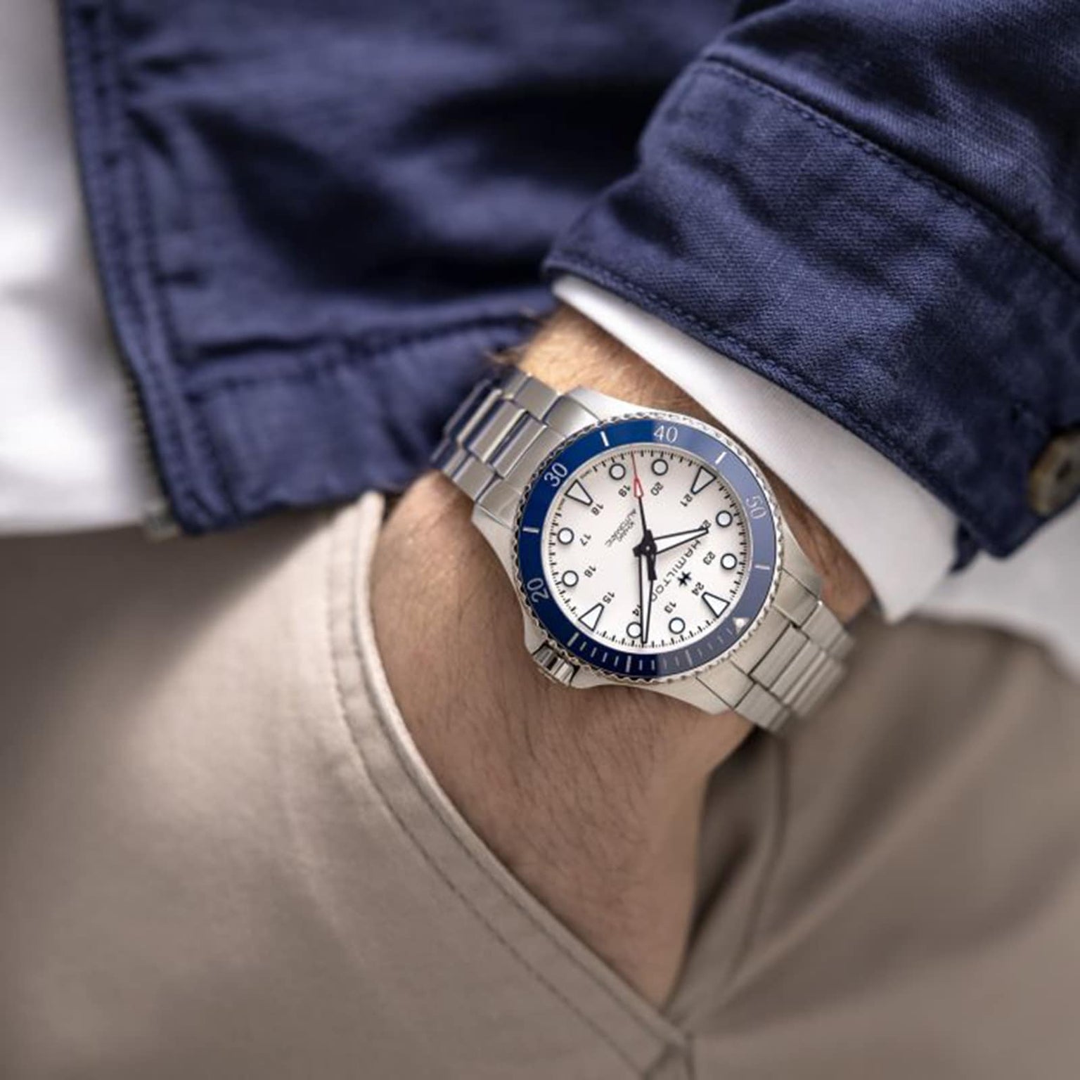 A person dressed in a blue jacket and beige pants sports the Hamilton Khaki Navy Scuba Auto, featuring a silver case, blue ceramic bezel, and white dial—perfect for a stylish summer at sea with their hand casually tucked in their pocket.