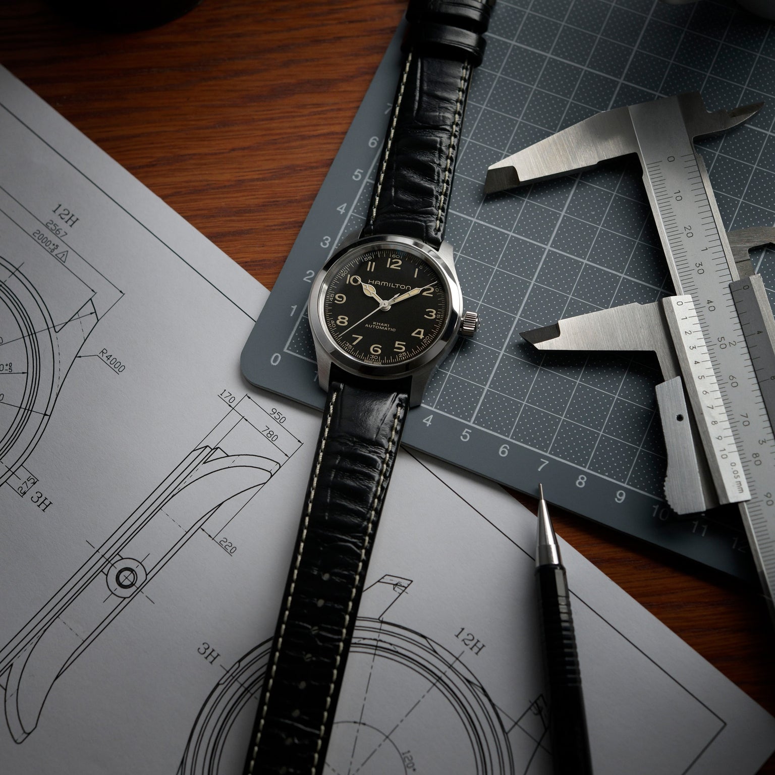 A Hamilton Khaki Field Murph 38mm with an 80-hour power reserve and black leather strap rests on a notebook with watch sketches, next to a caliper, ruler, and pen on a wooden desk.