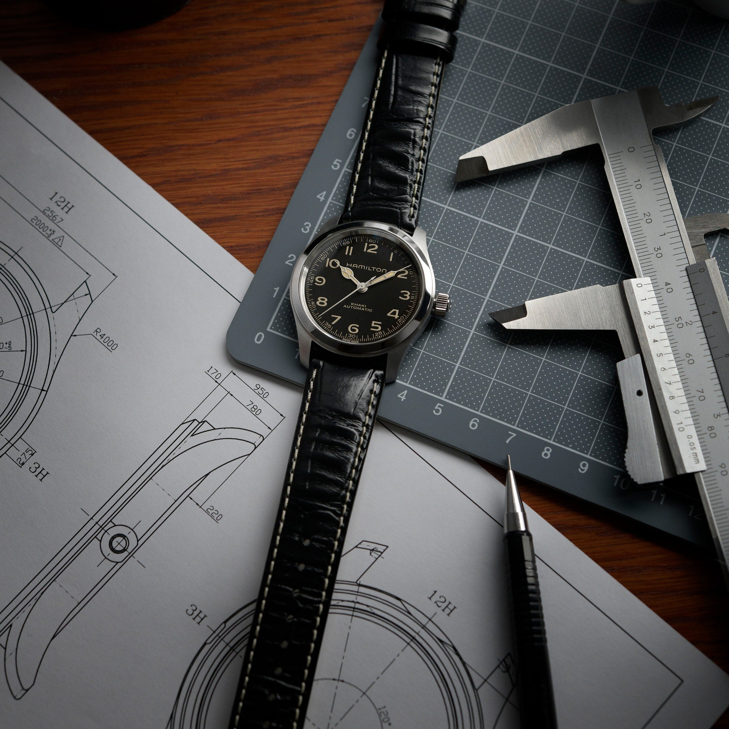 A Hamilton Khaki Field Murph 38mm with an 80-hour power reserve and black leather strap rests on a notebook with watch sketches, next to a caliper, ruler, and pen on a wooden desk.