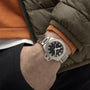 A person in a green jacket and orange sweater has their hand in their pocket, showing a Hamilton Khaki Navy Frogman Auto watch with a black face, silver metal band, and standout water resistance.