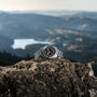 The Hamilton Khaki Field Expedition Auto, featuring a black dial and stainless steel case, rests atop a rocky surface with a blurred mountain landscape and lake in the background.