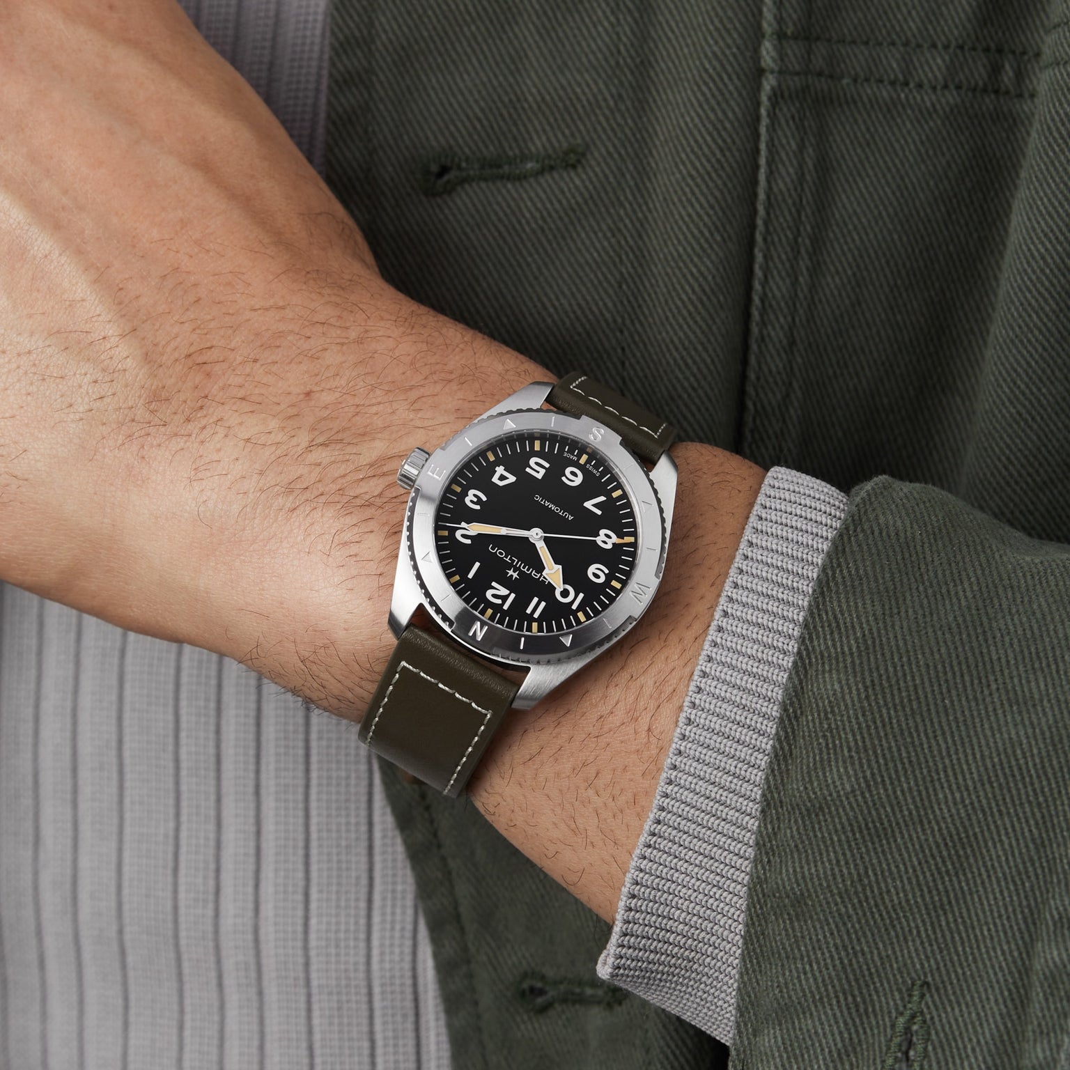 A person wearing the Hamilton Khaki Field Expedition Auto with a stainless steel case, black dial, white numerals, and an olive green leather strap, dressed in a green jacket and gray shirt.