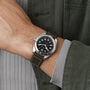 A person wearing the Hamilton Khaki Field Expedition Auto with a stainless steel case, black dial, white numerals, and an olive green leather strap, dressed in a green jacket and gray shirt.