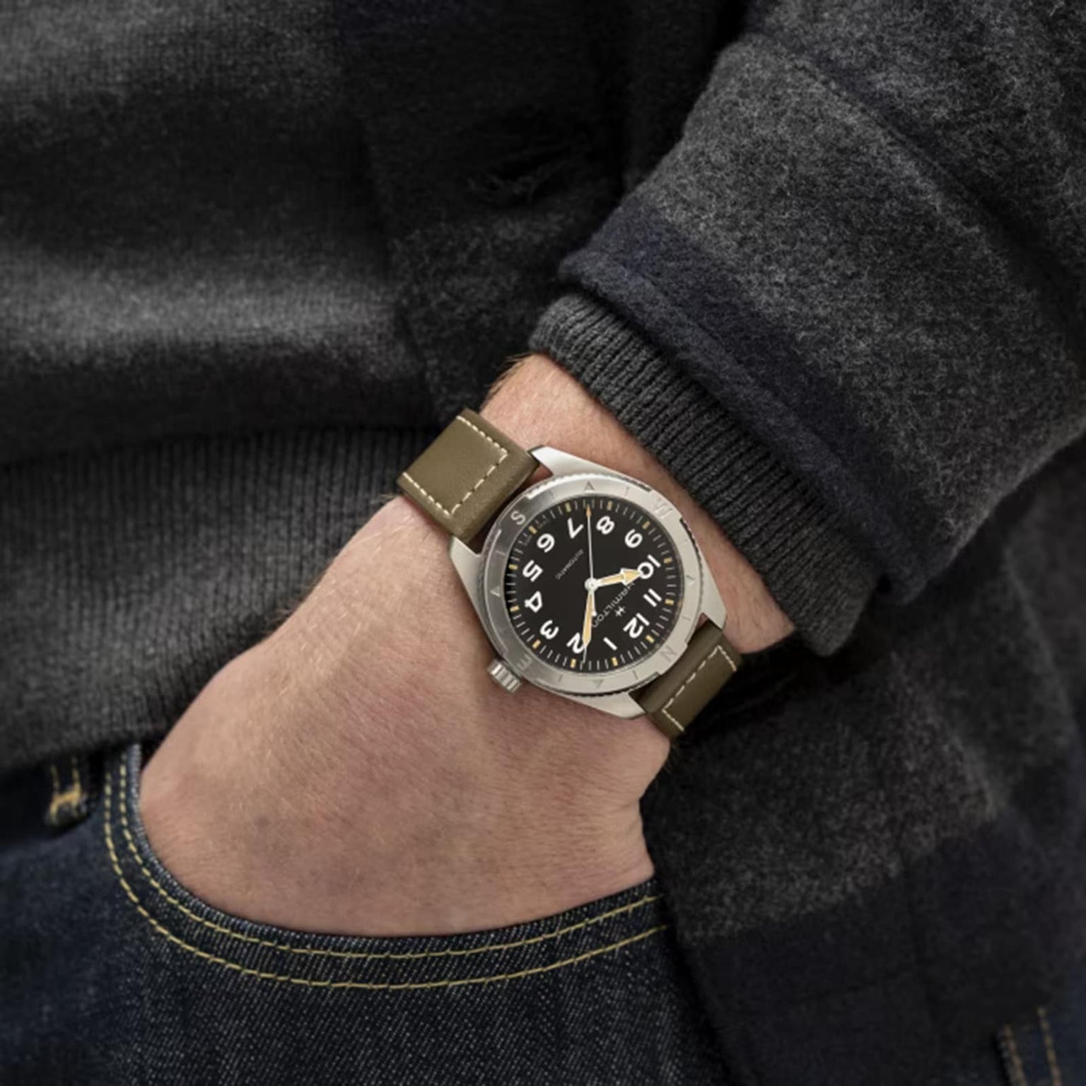 A person wearing the Hamilton Khaki Field Expedition Auto, a brown-strapped watch with stainless steel case and black dial, featuring Super-LumiNova® white numerals, has their hand partially in the pocket of blue jeans.