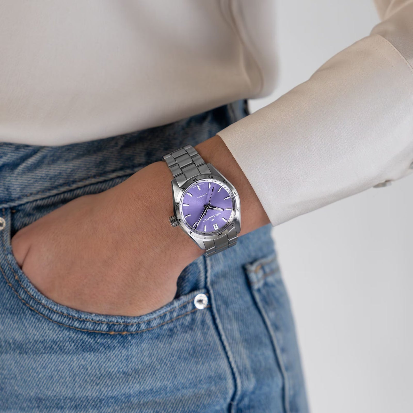 Wearing a cream shirt and blue jeans, a person with one hand in their pocket sports the Hamilton Jazzmaster Performer Auto—elegant with its stainless steel case and striking purple dial.