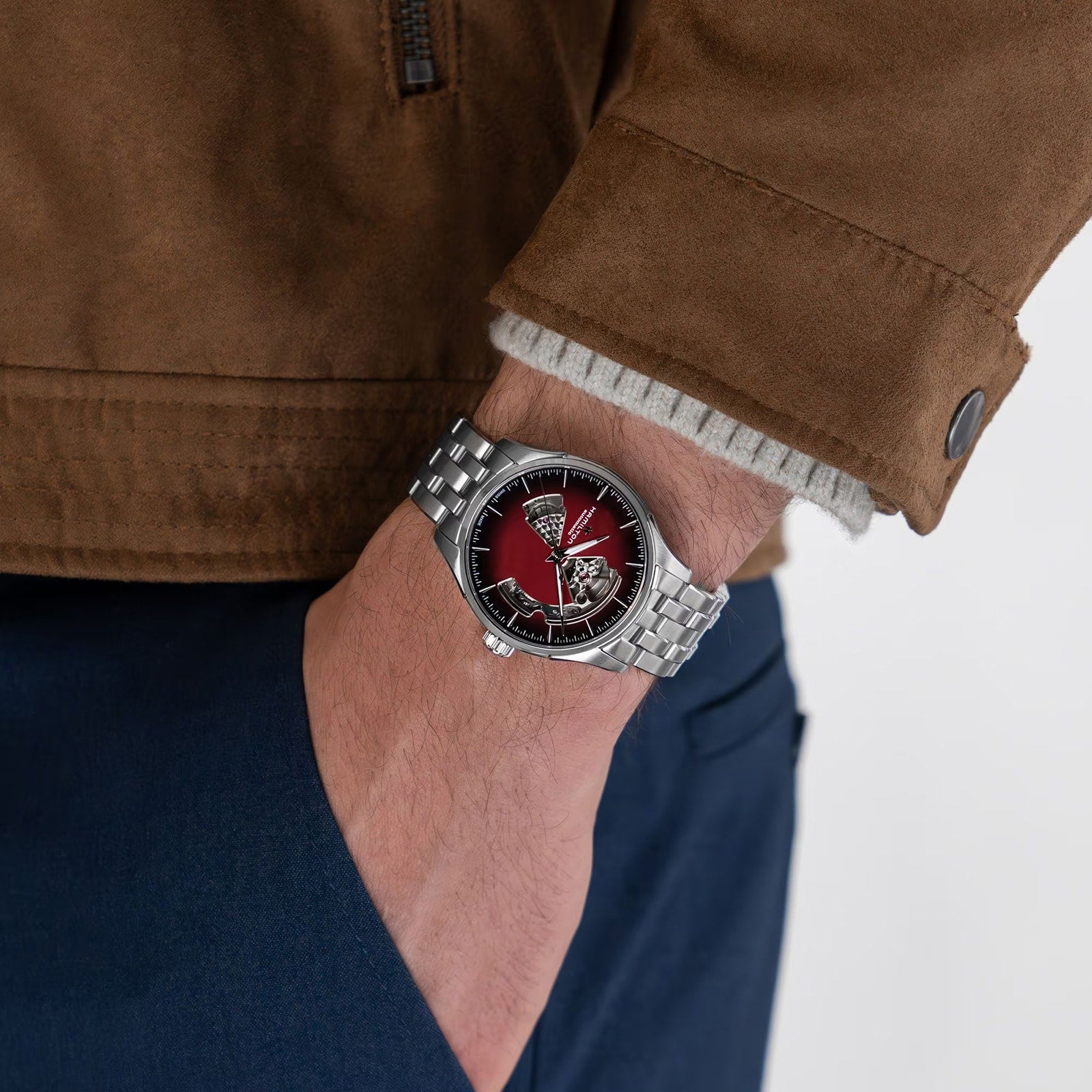 A person in a brown jacket and blue pants has their hand in their pocket, showing off a Hamilton Jazzmaster Open Heart Auto wristwatch with a red dial.