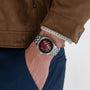 A person in a brown jacket and blue pants has their hand in their pocket, showing off a Hamilton Jazzmaster Open Heart Auto wristwatch with a red dial.