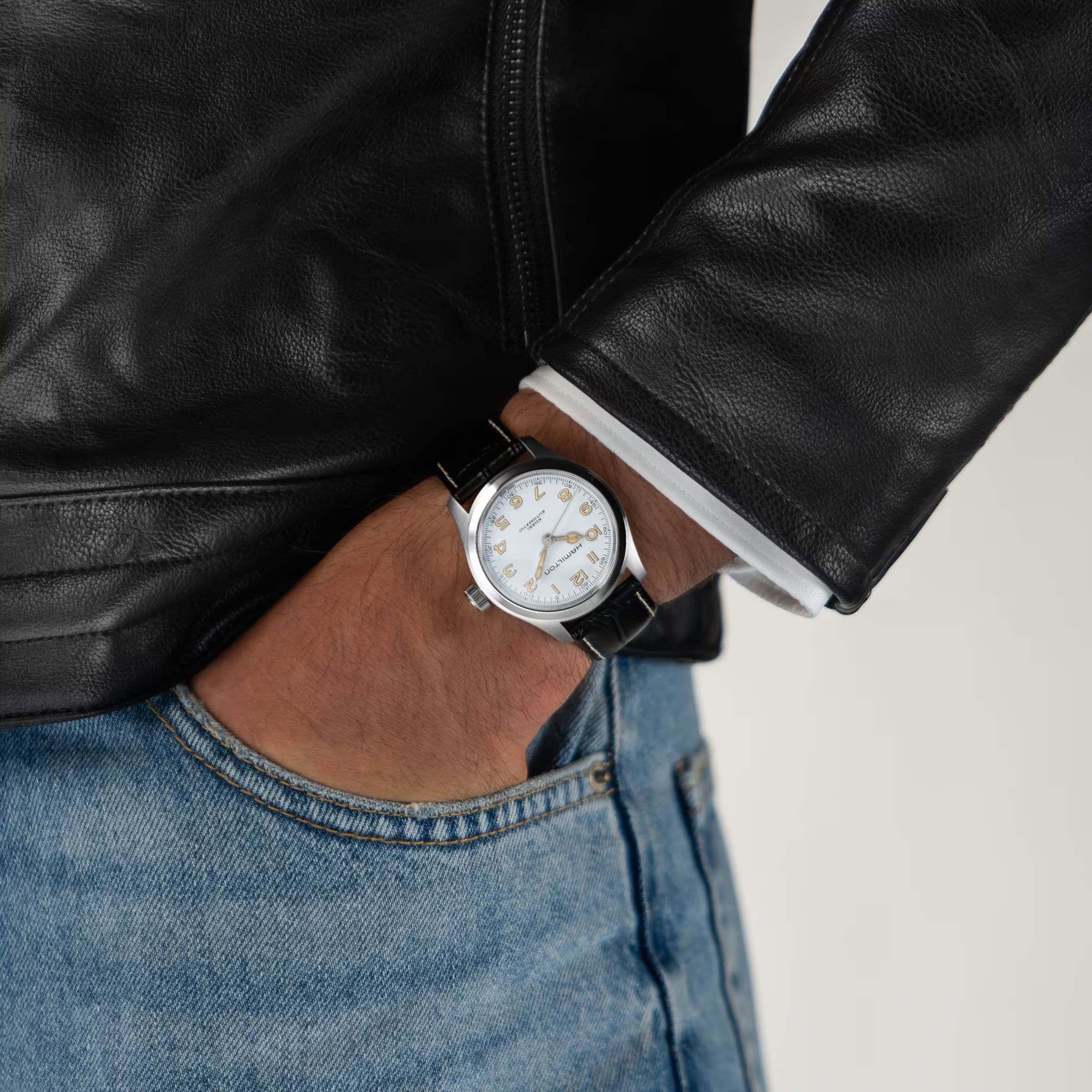 A person in a black leather jacket and blue jeans has their left hand in their pocket, showcasing the Hamilton Khaki Field Murph 38mm—a vintage-inspired timepiece with a white face and black leather strap beloved by enthusiasts.