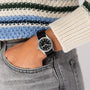 A person wears a Hamilton Khaki Field Quartz 33mm with a black round face and white numbers, paired with gray jeans and a striped knitted sweater, one hand in their pocket, reflecting subtle military heritage.