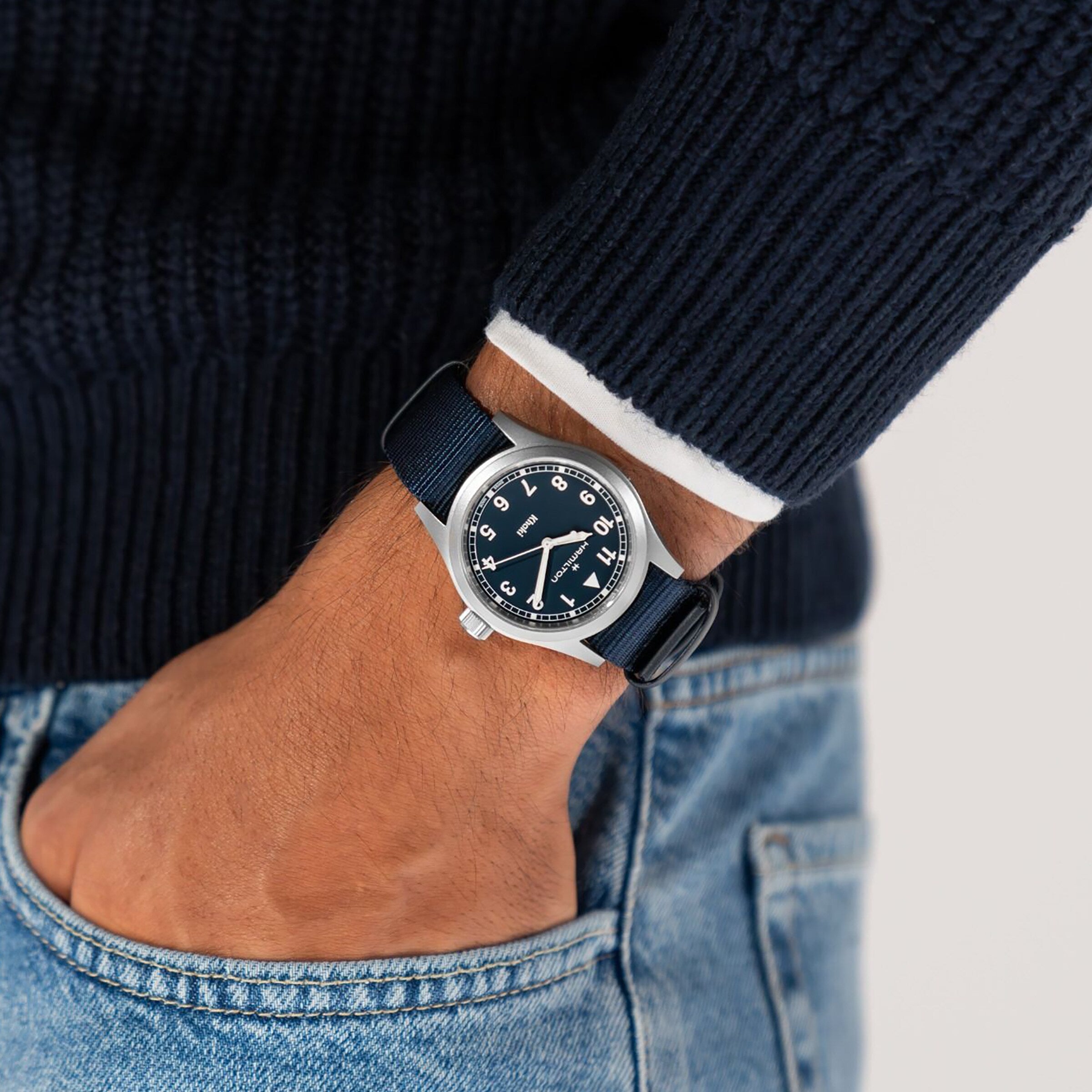 A person in a navy sweater and light blue jeans wears the Hamilton Khaki Field Quartz 38mm wristwatch with a dark blue dial and NATO strap, showcasing classic military heritage, on their left wrist.