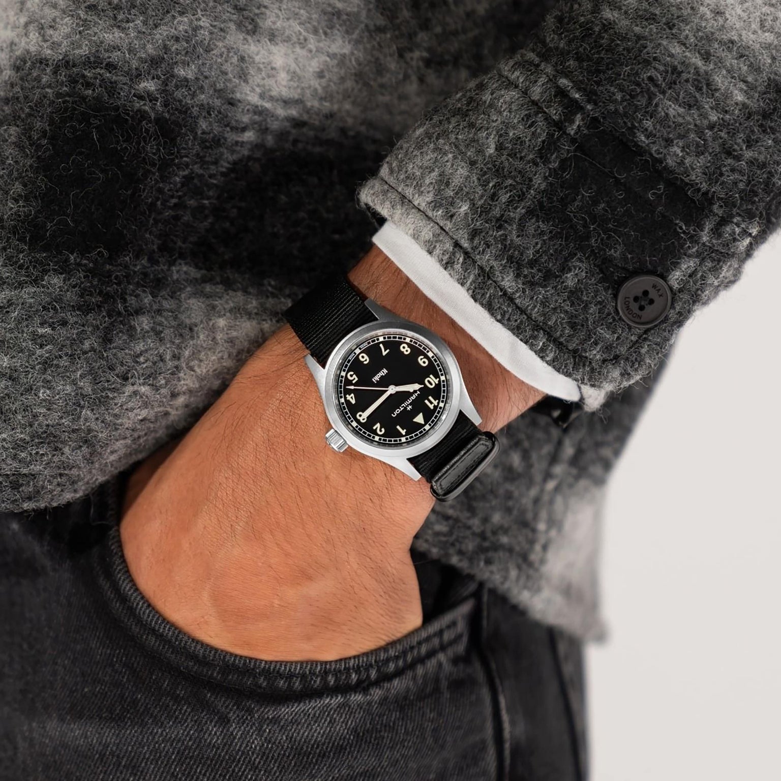 A person wearing a Hamilton Khaki Field Quartz watch with a black strap and a gray plaid jacket, hand in pocket.