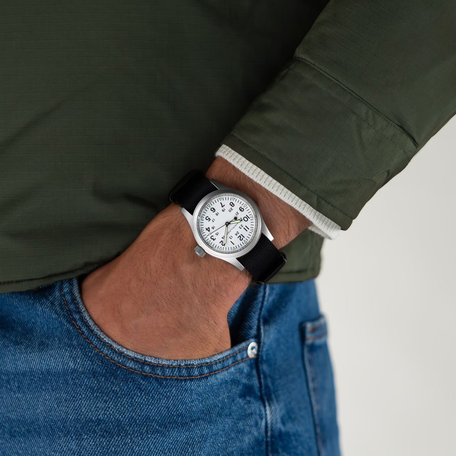 A person in a green jacket and blue jeans, with their left hand in their pocket, wears a Hamilton Khaki Field Mechanical watch featuring a white face and classic NATO strap.